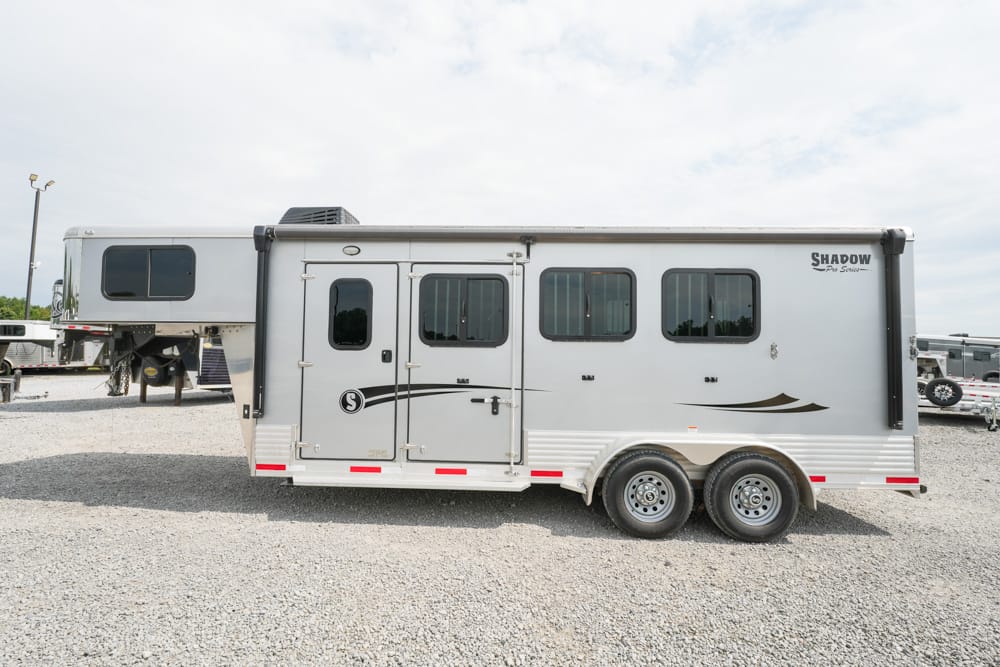 Silver horse trailer with living quarters and two axles parked on gravel lot.