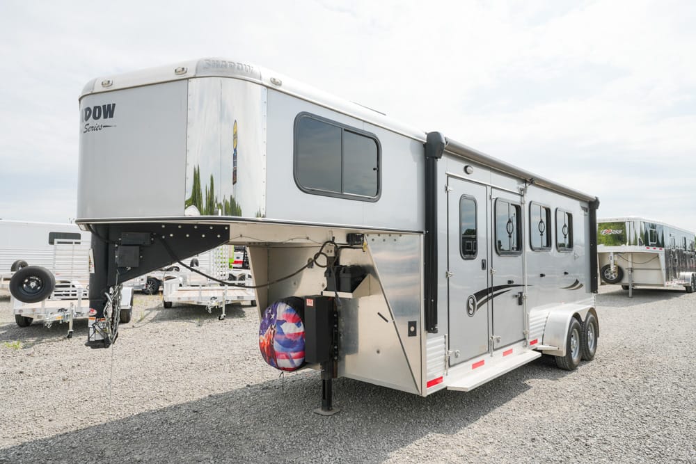 Silver gooseneck horse trailer with three axles, parked on gravel; spare tire cover features an American flag.