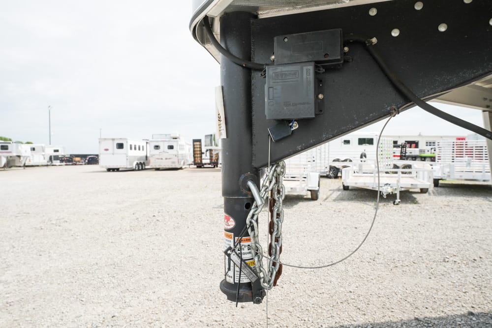 Close-up of a trailer hitch with safety chains and electrical plug on a gravel lot with horse trailers in the background.