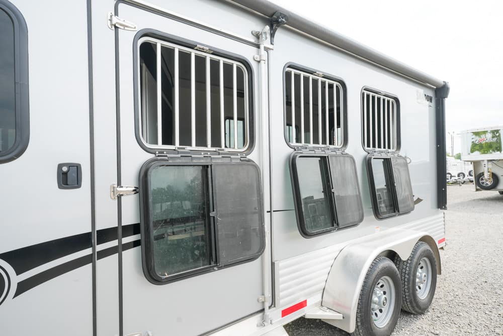 Gray horse trailer with barred windows and dual wheels parked on a gravel lot.