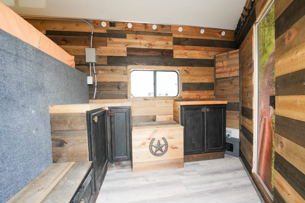 Wood-paneled tiny kitchen inside a trailer with black cabinets, a central storage chest, and a window above the counter
