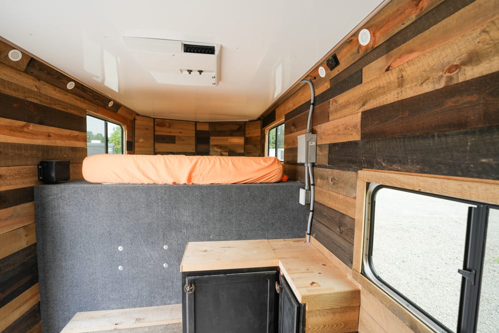 Interior of a rustic tiny camper: bed on a gray platform with orange bedding, wood plank walls, and a window on the side.