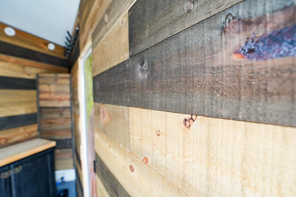 Close-up of weathered wooden planks forming a wall inside a shed, with a doorway and a shelf on the left.