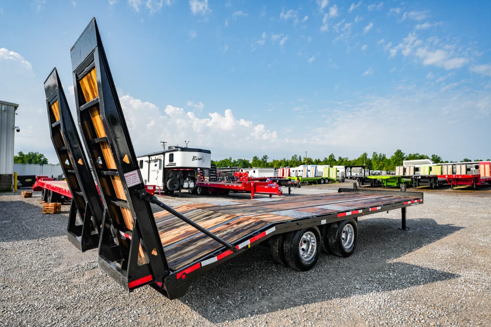 Flatbed trailer with raised rear ramps in a gravel lot, with colorful trailers in the background under a blue sky