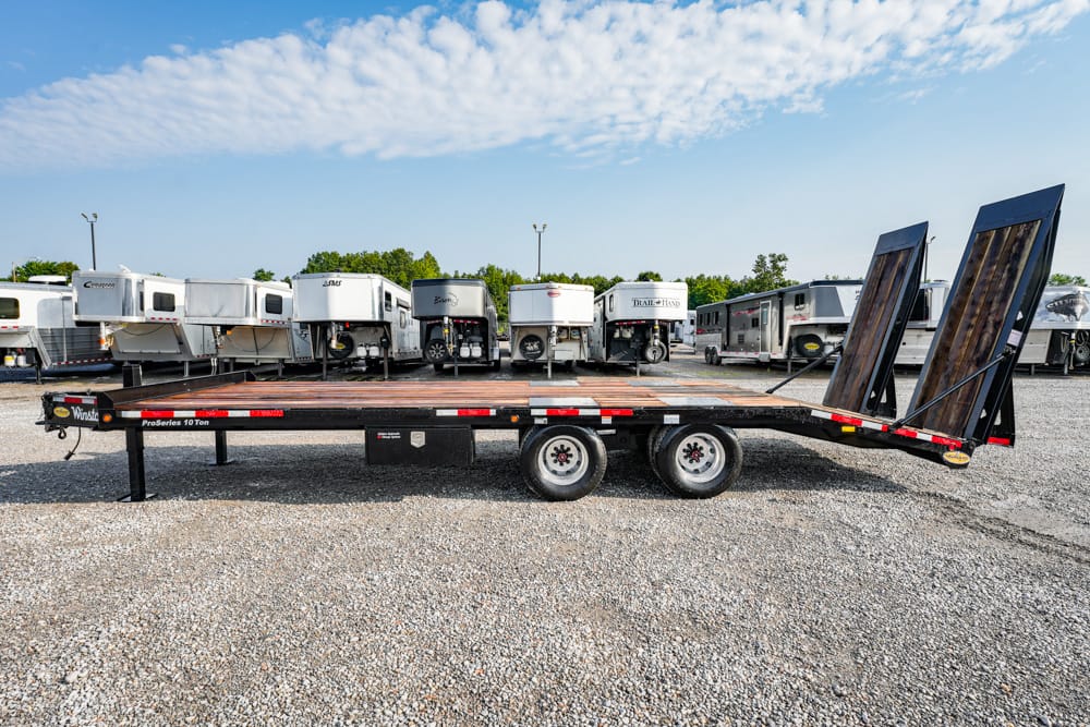 Flatbed trailer with dual ramps raised at the rear, parked in a gravel lot with RVs in the background (trailer for hauling vehicles).
