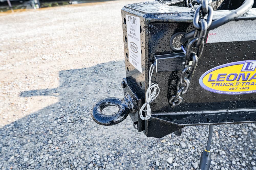 Close-up of a black trailer hitch with a circular hitch ring, chains, and a Leonard Truck & Trailer sticker on a wet gravel surface.
