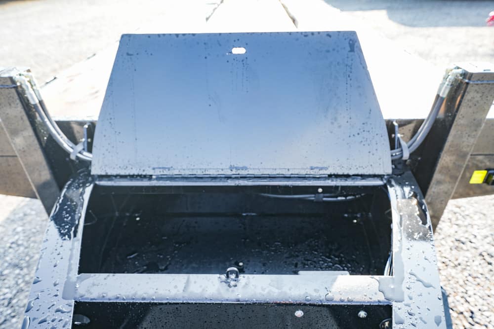 Open metal hopper with its lid raised, showing a dark wet interior and water droplets on the exterior outdoors on a gravel surface.