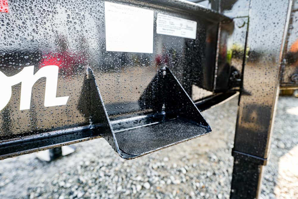 Close-up of a black metal trailer frame with a folded step, wet with rain droplets on a gravel lot.