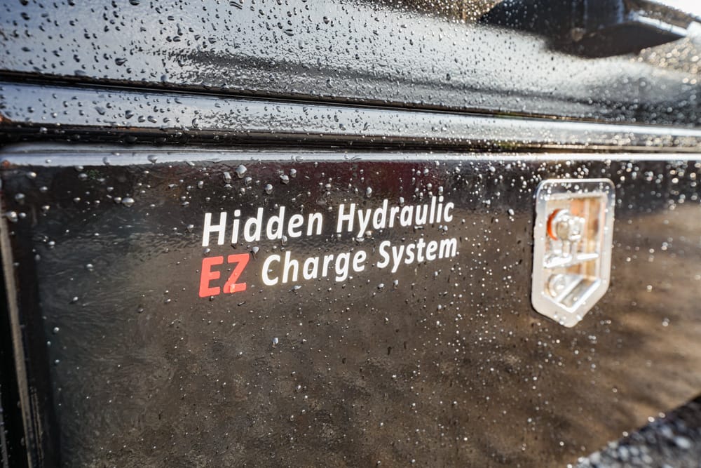 Close-up of a black, rain-speckled vehicle panel showing the label 'Hidden Hydraulic EZ Charge System' beside a metal charger port.