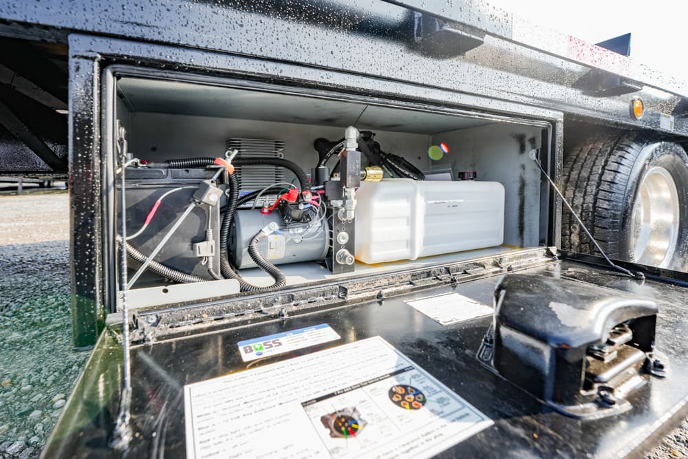 Open service compartment on a truck trailer showing a white plastic tank, battery, and various hoses and wiring inside the bay.