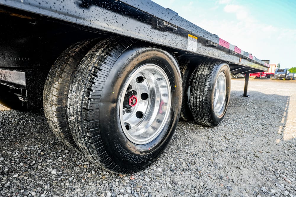 Close-up of dual trailer wheels with chrome rims on a gravel lot, part of a black flatbed trailer underside visible above the tires.