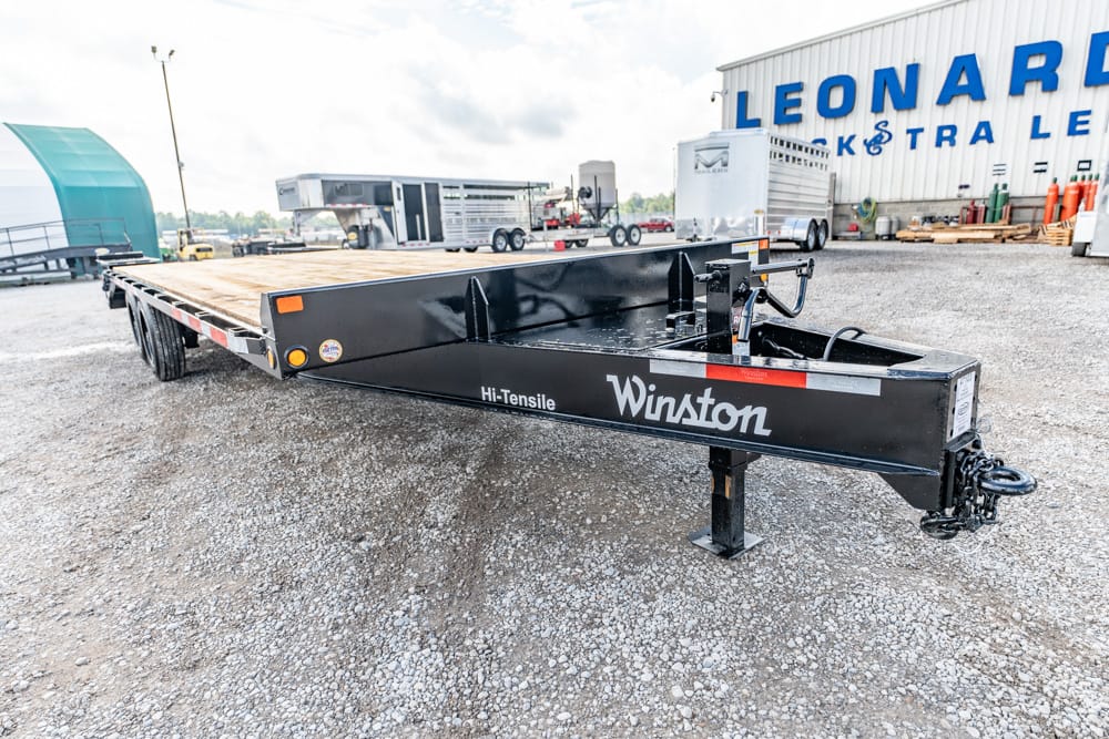 Black Winston Hi-Tensile flatbed trailer with wooden deck sits on a gravel lot in front of a building with blue signage (LEONARD).