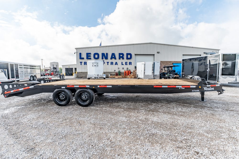Long flatbed trailer with dual rear axles parked on a gravel lot in front of a building with blue signage.