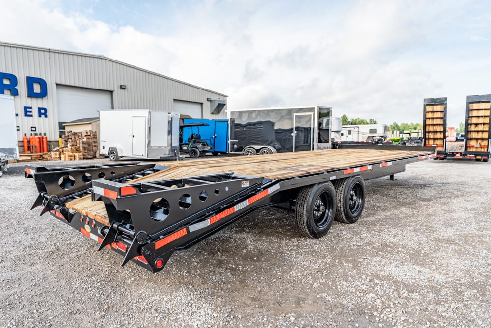 Long flatbed trailer with a wooden deck parked in a gravel lot near an industrial building.