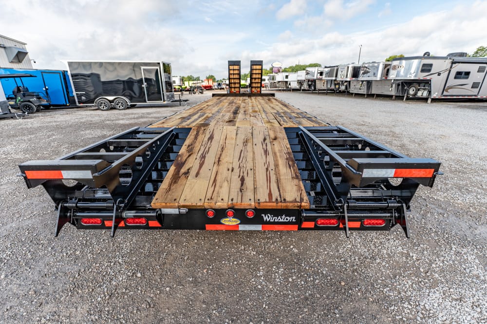 Long flatbed trailer with a wooden deck and black metal frame parked in a lot of trailers on gravel ground.
