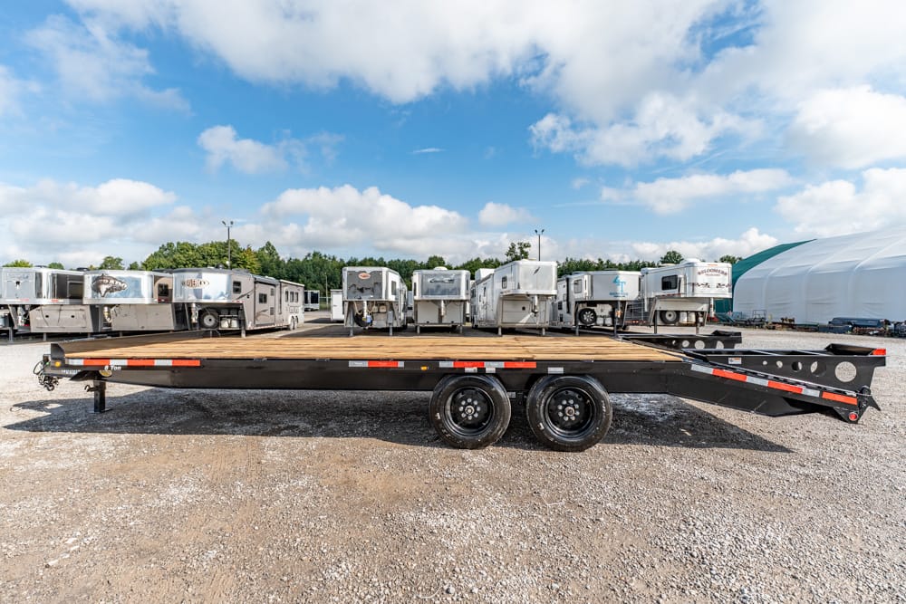 Black flatbed trailer with dual rear wheels on a gravel lot, rows of travel trailers behind it and a large white tent in the distance.