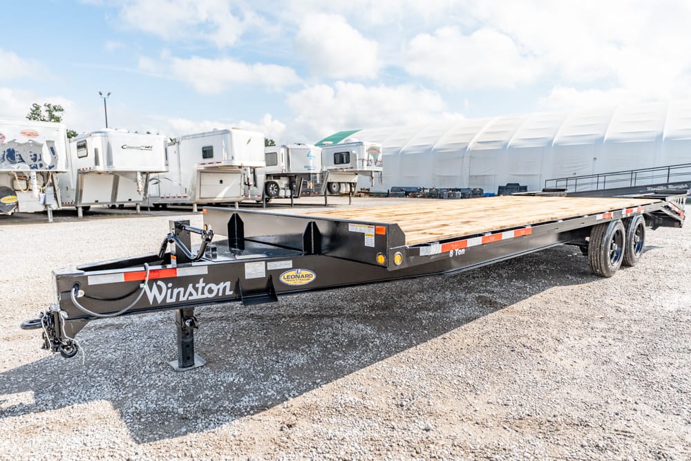 Black Winston flatbed trailer with a wooden deck, 8-ton capacity, parked on a gravel lot with other trailers in the background.
