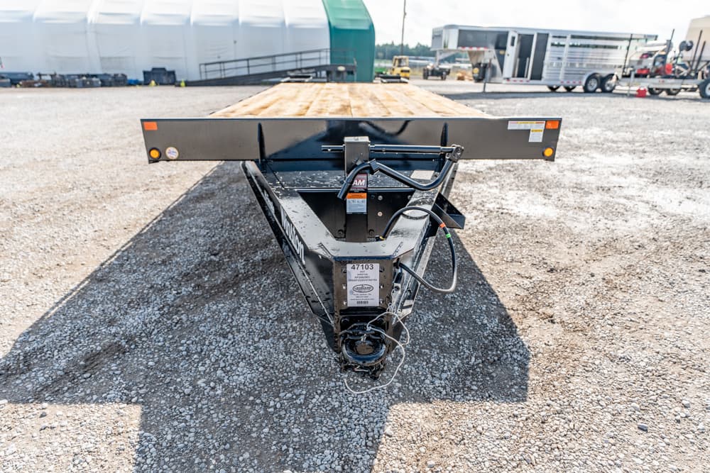 Front view of a black equipment trailer with a wooden flatbed parked on gravel, industrial gear in the background.
