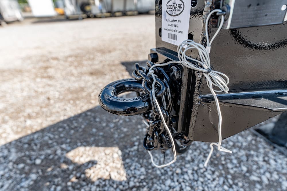 Close-up of a trailer hitch with a circular coupler, tangled white wires, and a tag on a gravel lot.