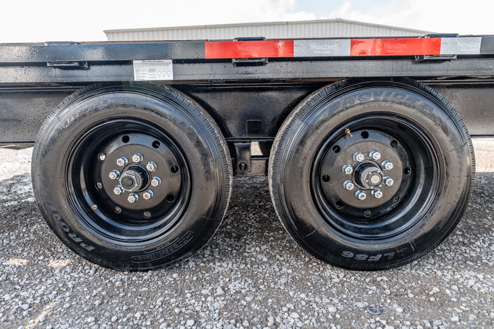 Two black trailer wheels with lug nuts on a gravel surface under a flatbed trailer (dual axle)