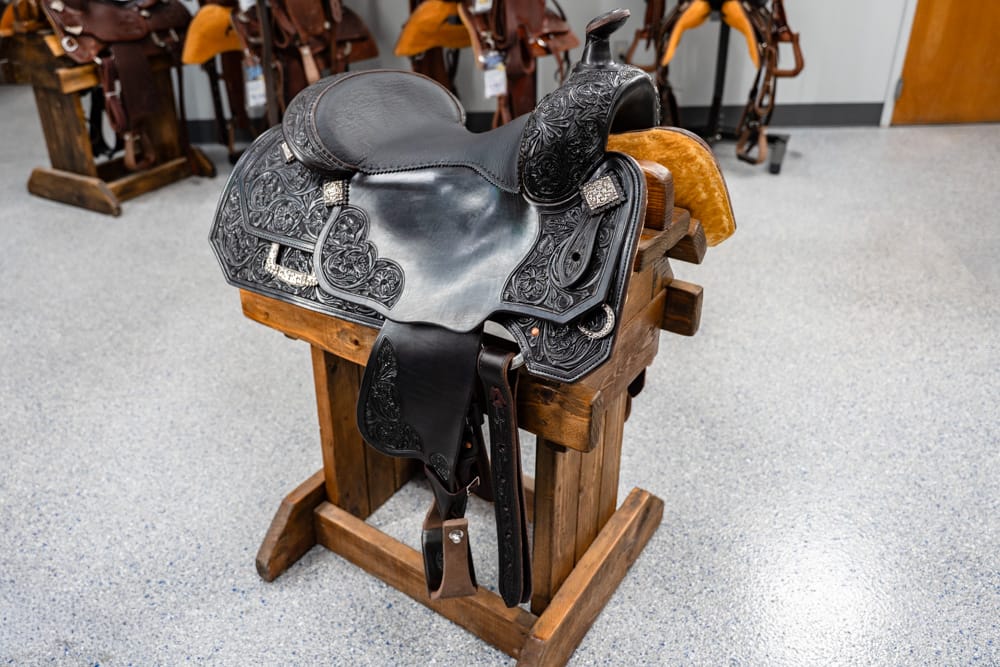 Black tooled Western saddle with ornate floral pattern on a wooden stand in a tack room/showroom with other saddles in the background