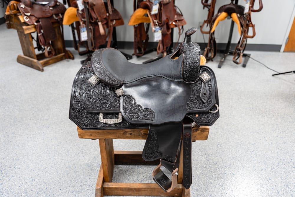Black leather Western saddle with intricate floral tooling, displayed on a wooden stand in a tack shop.