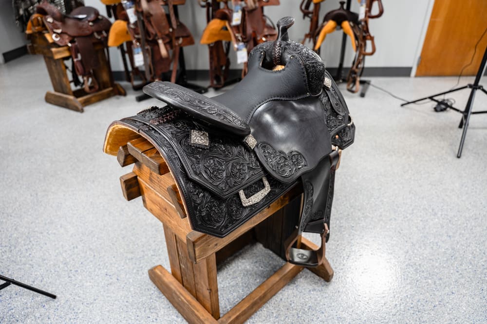 Black tooled leather western saddle on a wooden stand in a tack shop with other saddles in the background.