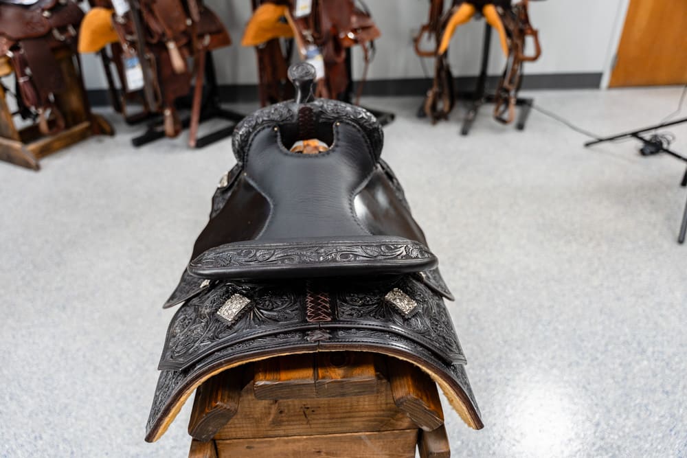 Close view of a intricately tooled black leather western saddle on a wooden rack, with other saddles in the background.