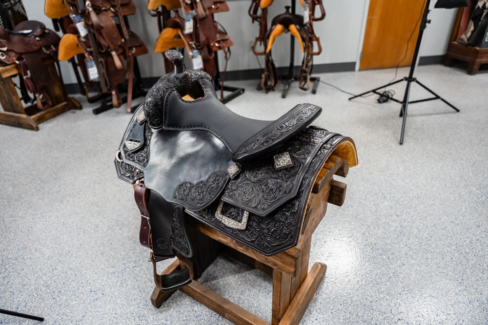 Ornate black Western saddle on a wooden rack in a tack shop, with more saddles in the background.