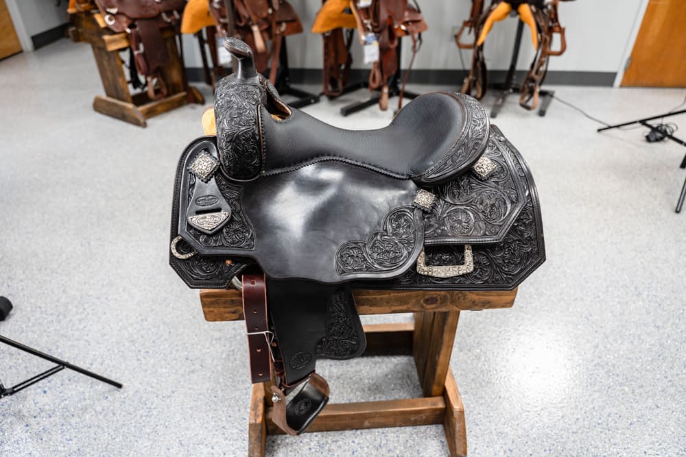 Ornate black Western saddle with floral tooling on a wooden rack in a tack room showroom.