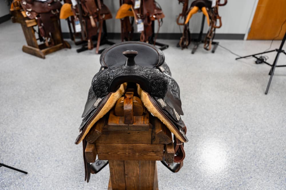 Western saddle on a wooden stand with other saddles blurred in the background, in a tack shop or classroom setting