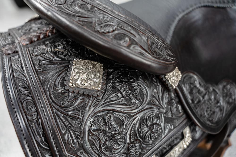 Close-up of a dark tooled leather saddle with floral engravings and decorative metal conchos.