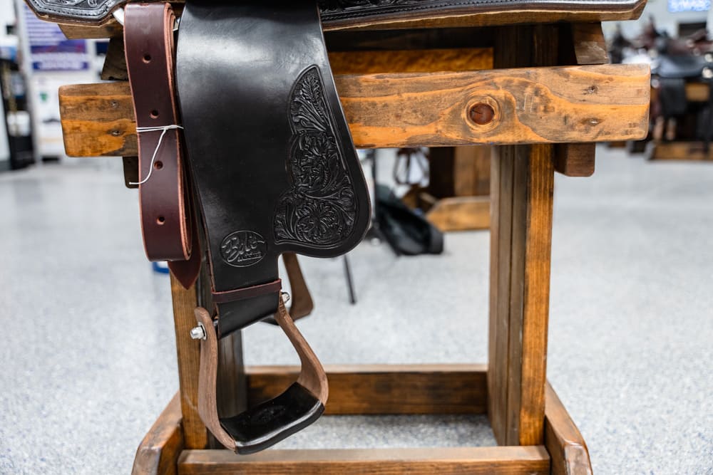 Leather western saddle with ornate tooling hanging on a wooden saddle rack in a shop display, stirrup strap visible on the left.