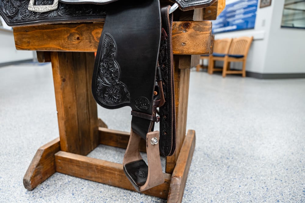 Leather Western saddle with decorative tooling hanging on a wooden saddle stand in an indoor room with chairs in the background