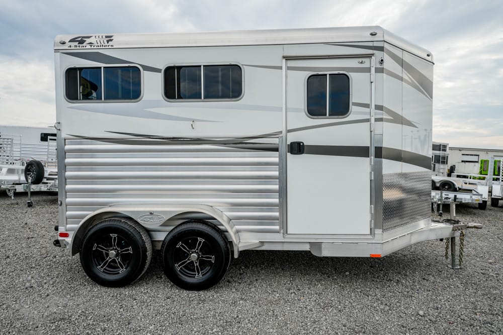 Silver aluminum horse trailer with two axles and black-rimmed wheels parked on a gravel lot, featuring side windows and a main entry door on the side panel.