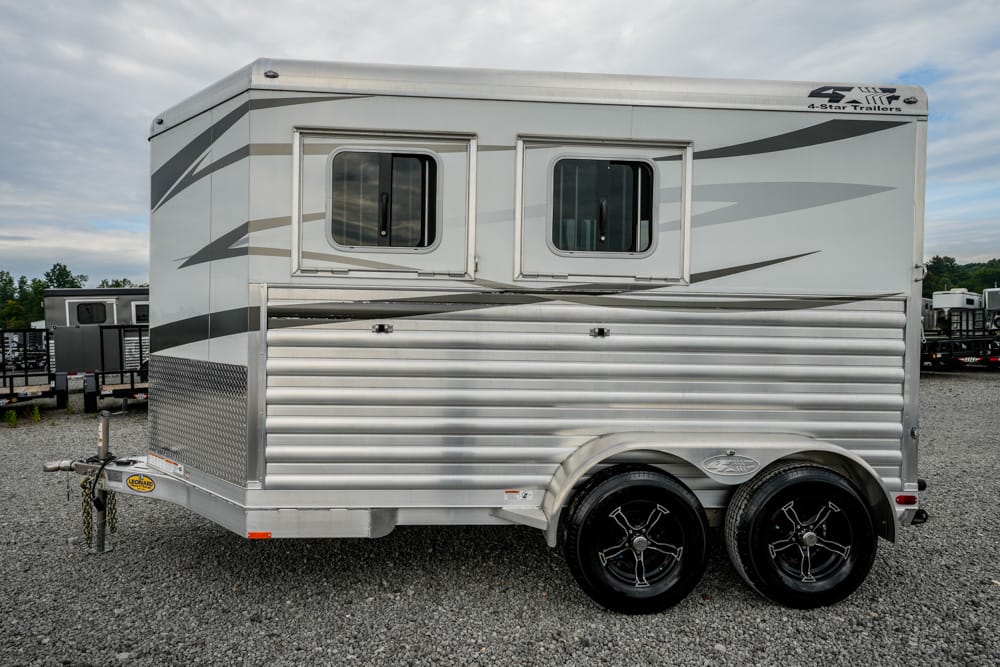 Silver aluminum travel trailer with two small windows and twin-axle wheels parked on gravel, showing side and front hitch area.