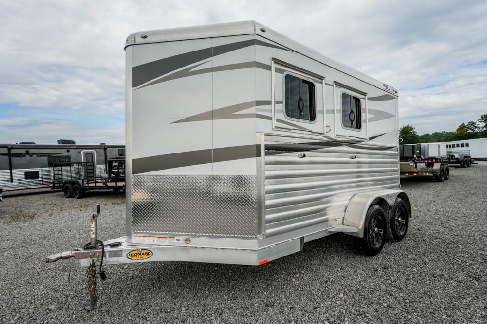 Silver travel trailer with two windows and diamond-plate front, parked on a gravel lot under a cloudy sky.