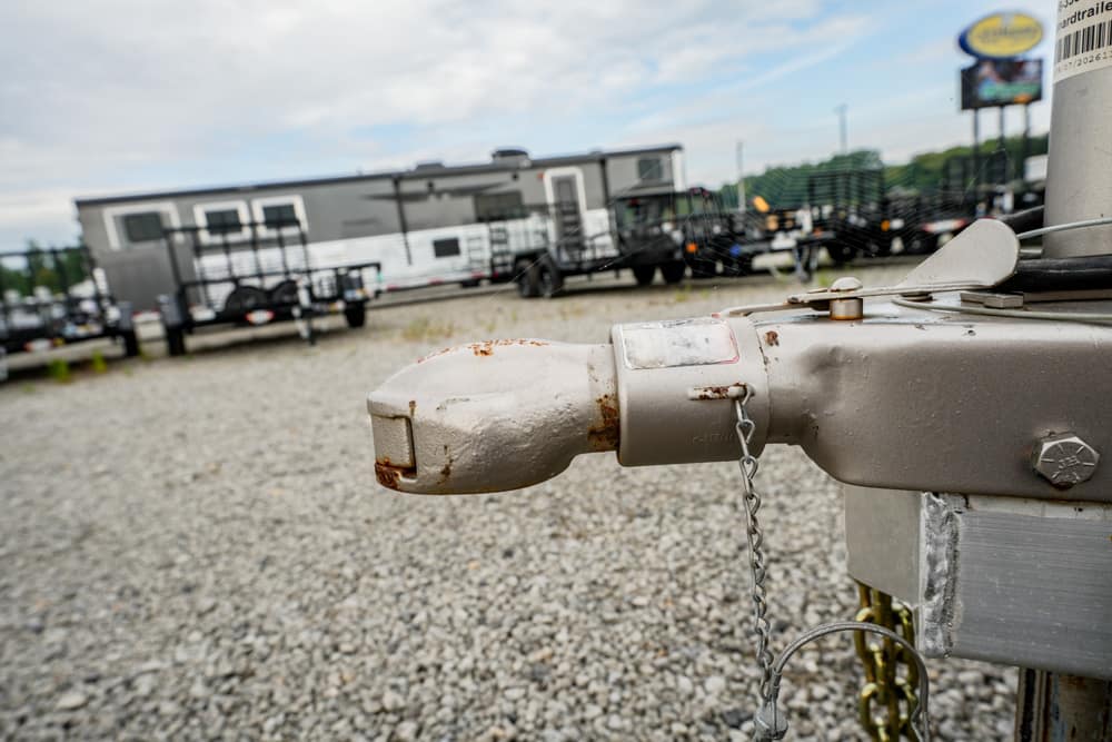 Close-up of a rusty trailer hitch in the foreground with a gravel lot and parked travel trailers in the background.