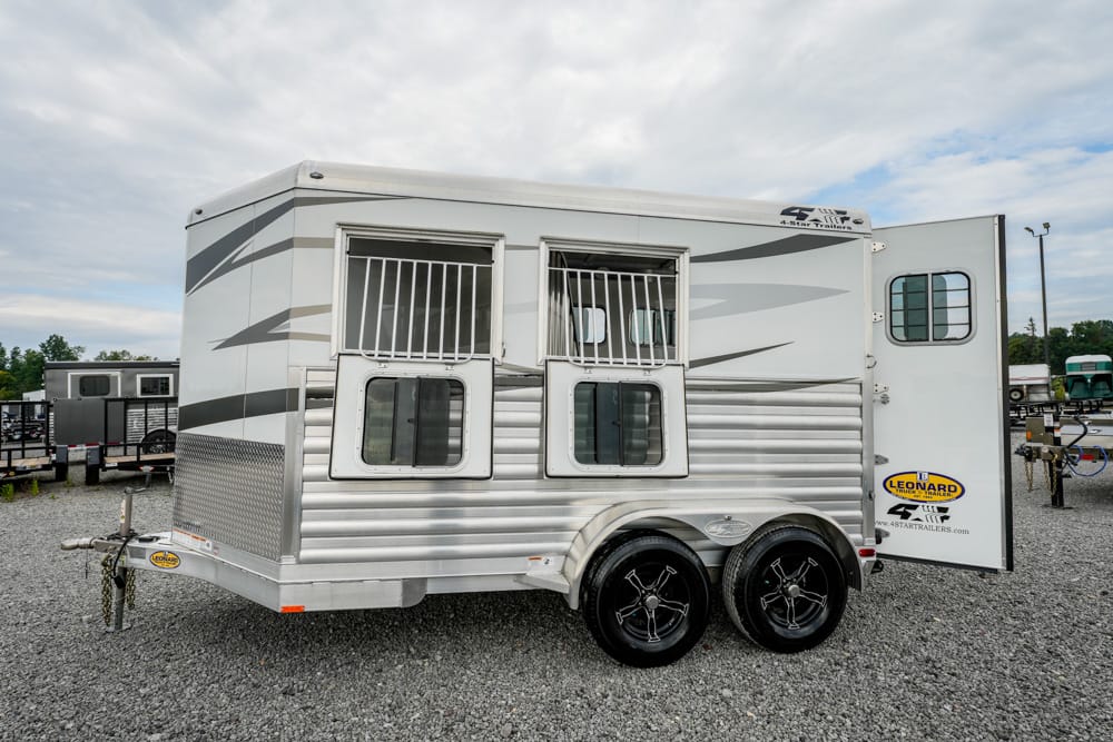 Aluminum horse trailer with two barred stall windows, open side door, and tandem wheels on a gravel lot.