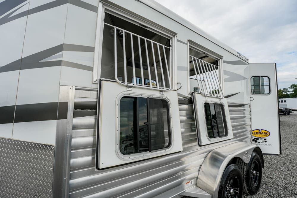 Silver aluminum horse trailer with two side windows, metal bars, and diamond-plate trim, parked on gravel.