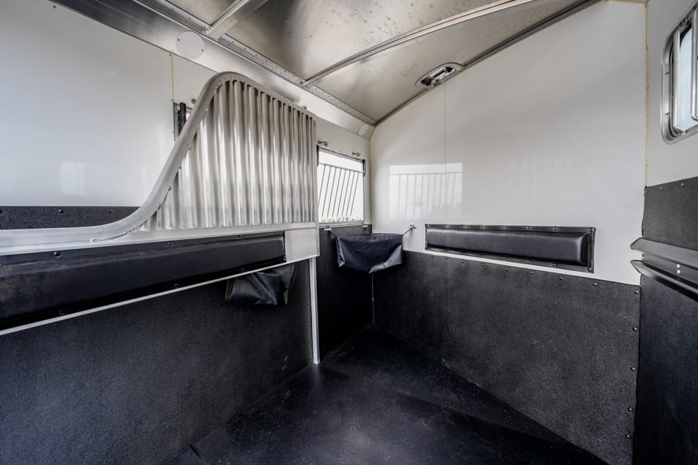 Interior of a horse trailer stall with a padded black divider, white walls, and a metal partition forming a curved gate nearby.