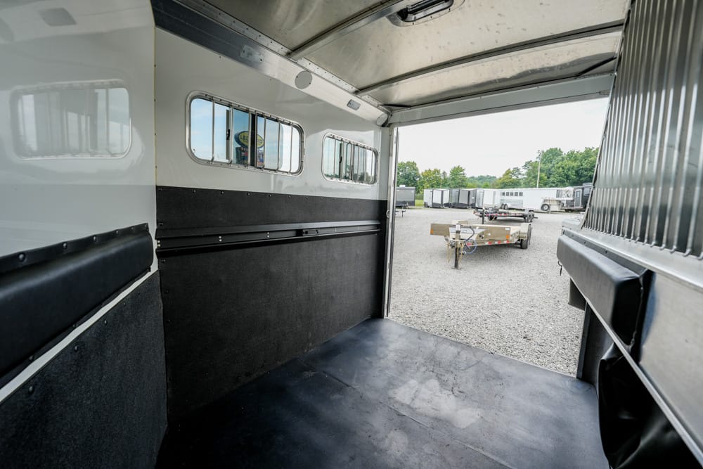 Interior of a white horse trailer with black padded walls and rubber floor, open to a gravel lot with parked trailers visible outside at the rear doorway.