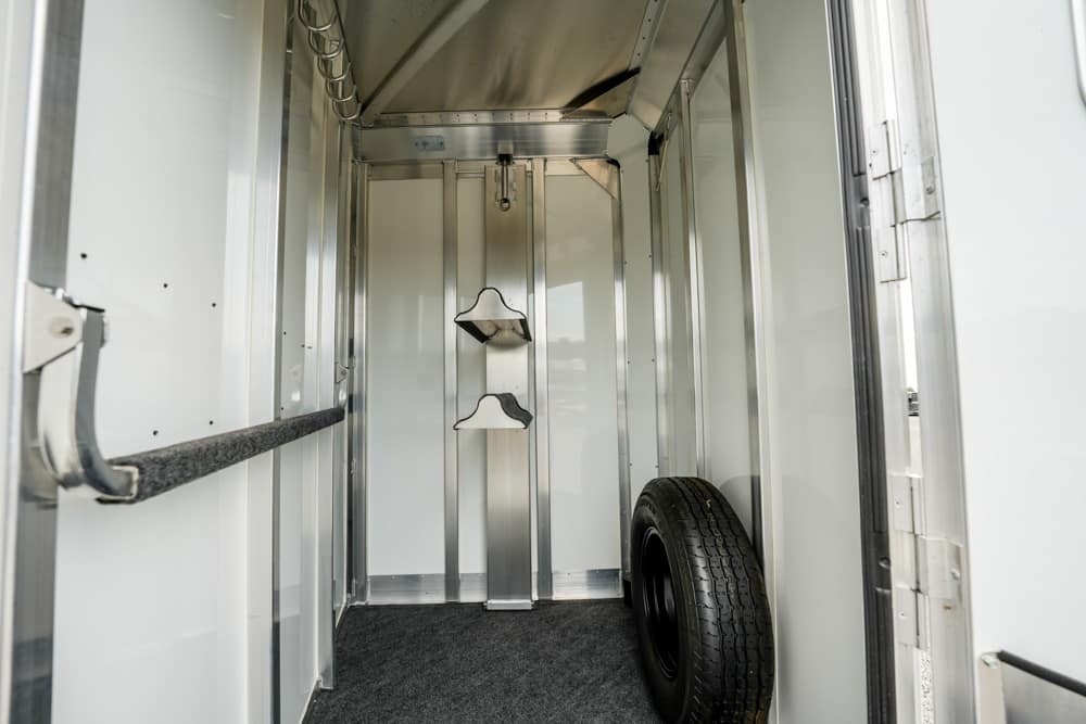 Interior of an empty metal horse trailer with two saddle racks on the back wall and a spare tire propped on the right side, finished with a carpeted floor.