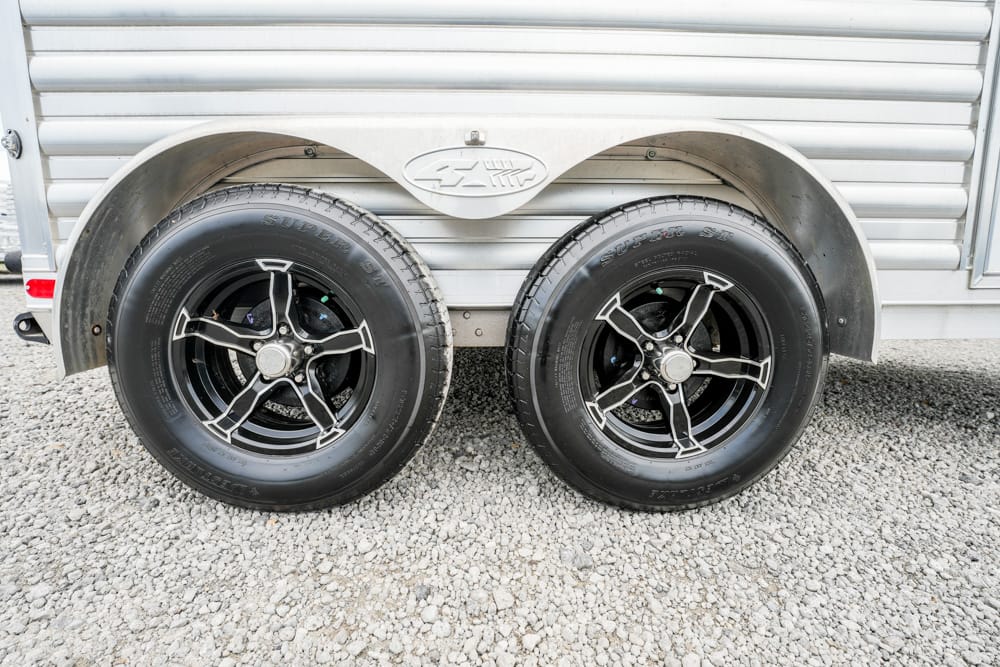 Dual trailer wheels with black rims on a silver cargo trailer, parked on gravel.