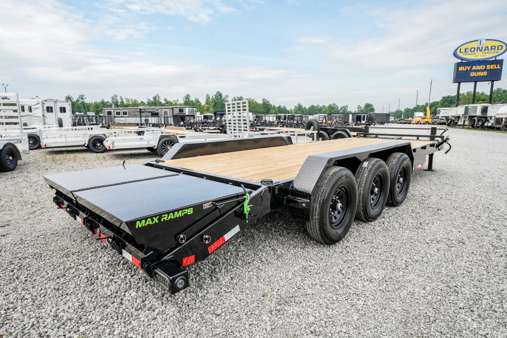 Flatbed trailer with wooden deck and three axles in a gravel lot at a trailer dealership, labeled MAX RAMPS on the side trench.