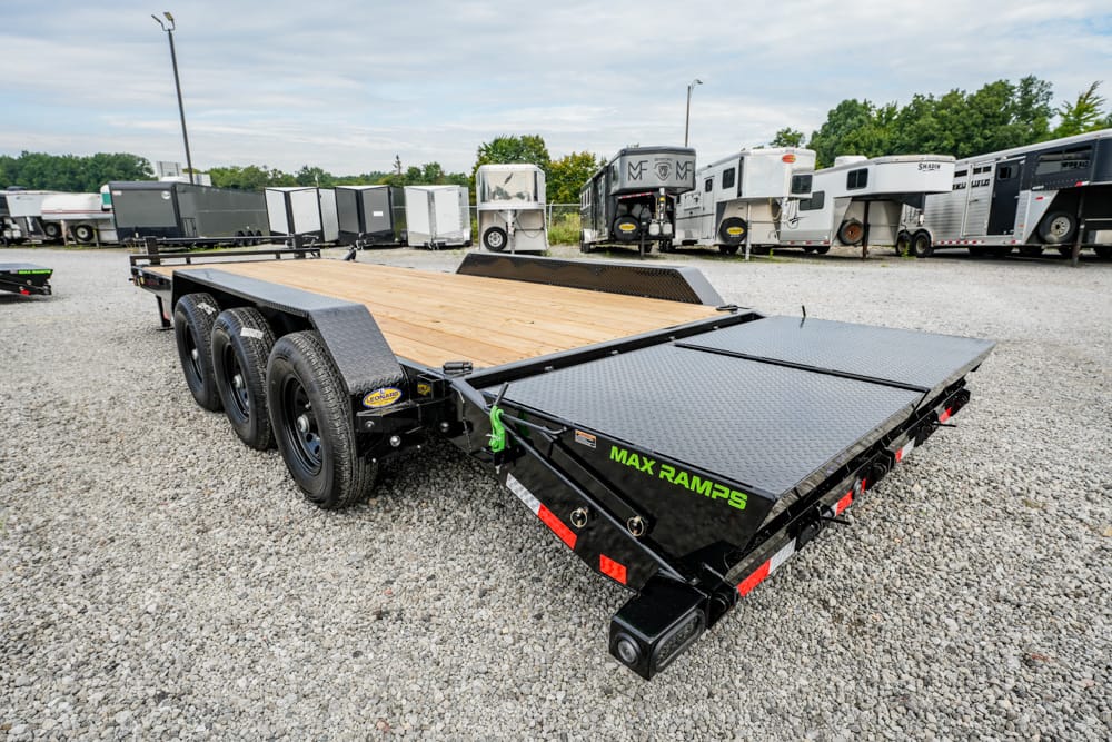 Flatbed trailer with a wood deck and diamond-plate rear ramps labeled MAX RAMPS in a gravel lot.