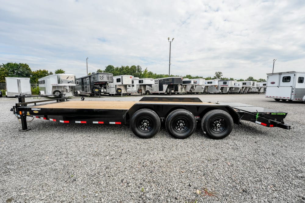 Black flatbed trailer with a wooden deck and three axles parked on gravel, with horse trailers in the background.
