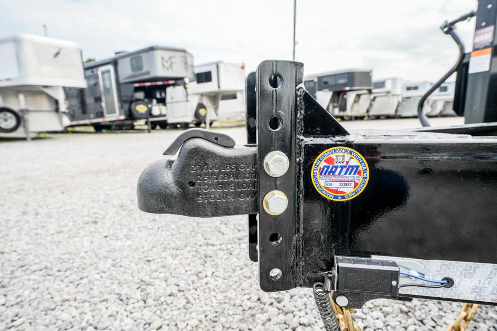 Close-up of a trailer hitch with a black coupler and bolts, attached to a black frame; gravel lot and travel trailers visible in the background.