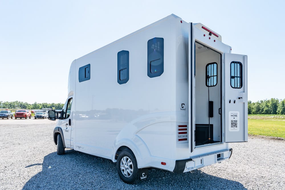 White recreational vehicle with rear entry door open, showing stairs inside on a gravel lot.