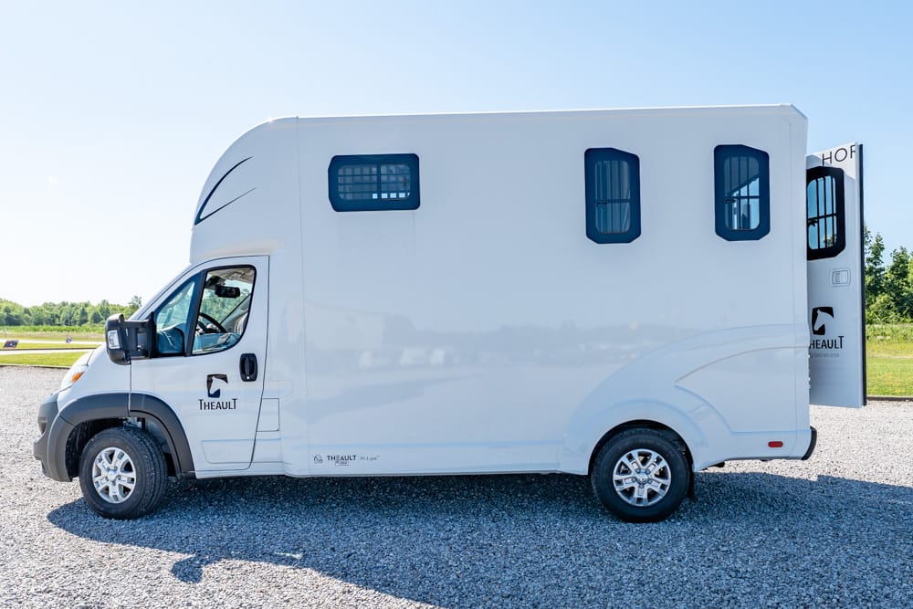 White motorhome (recreational vehicle) parked on a gravel lot, side view with its rear entry extended, blue-tinted windows and Theault branding visible against a clear blue sky.